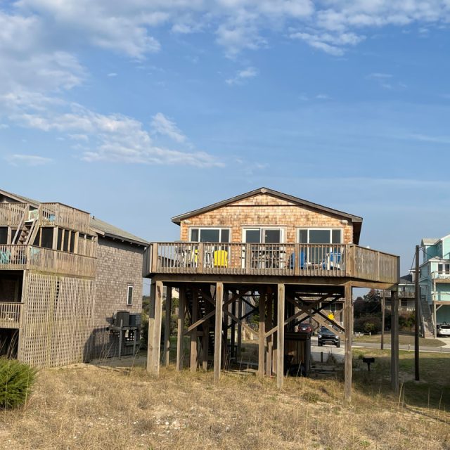 A wooden beach house on stilts with a large deck, surrounded by similar houses under a partly cloudy sky.