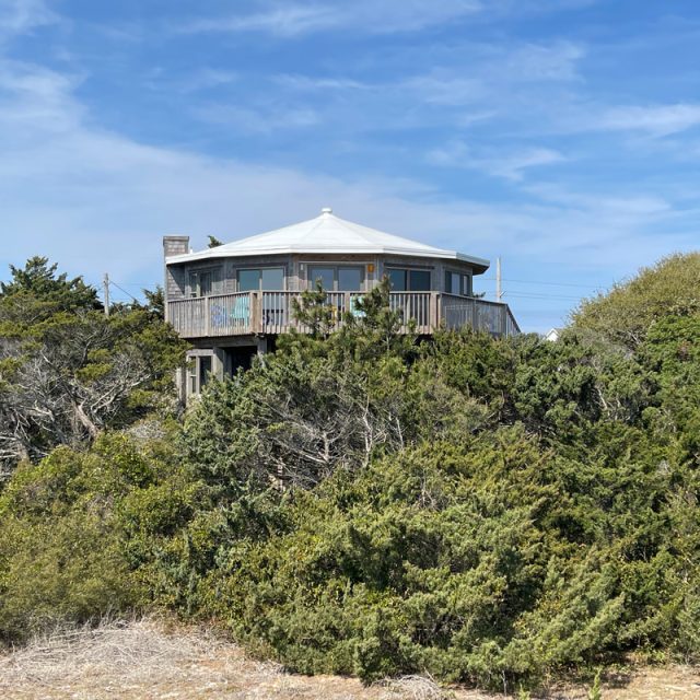 A round house with a wraparound deck is partially hidden by dense green shrubs under a blue sky.