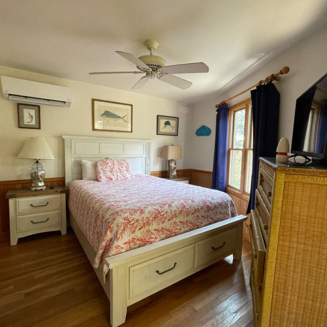 A bedroom with a double bed, nightstand, dresser, ceiling fan, window, and wall-mounted TV.