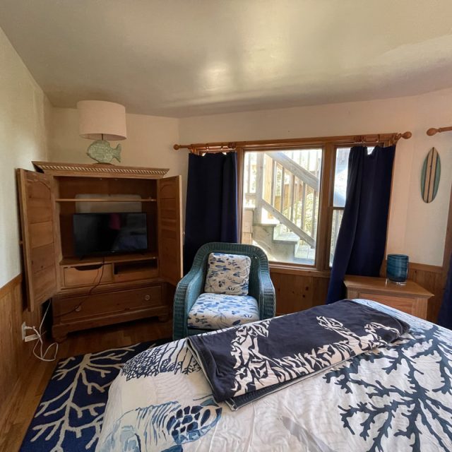 Cozy bedroom with a bed, TV in wooden cabinet, blue chair, and window showing wooden stairs outside.