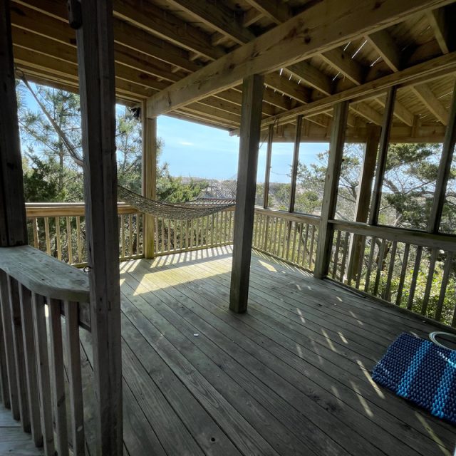 Wooden porch with a hammock, stairs, and trees outside. Blue mat and pet bowl on the deck floor in sunlight.