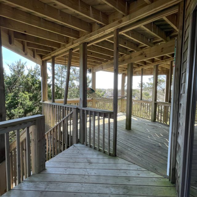 Wooden deck with railings and a roof, surrounded by trees, with sunlight streaming through the open sides.
