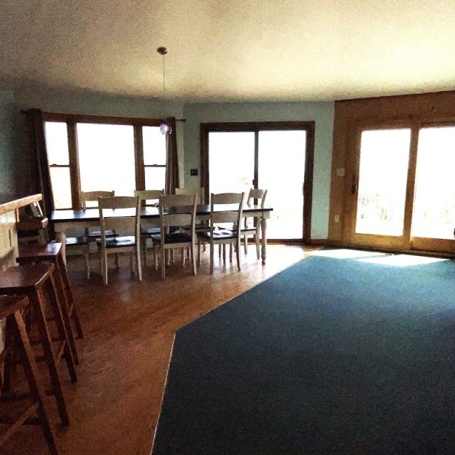 Sunlit dining area with a wooden table, white chairs, bar stools, and large windows by a blue carpeted floor.