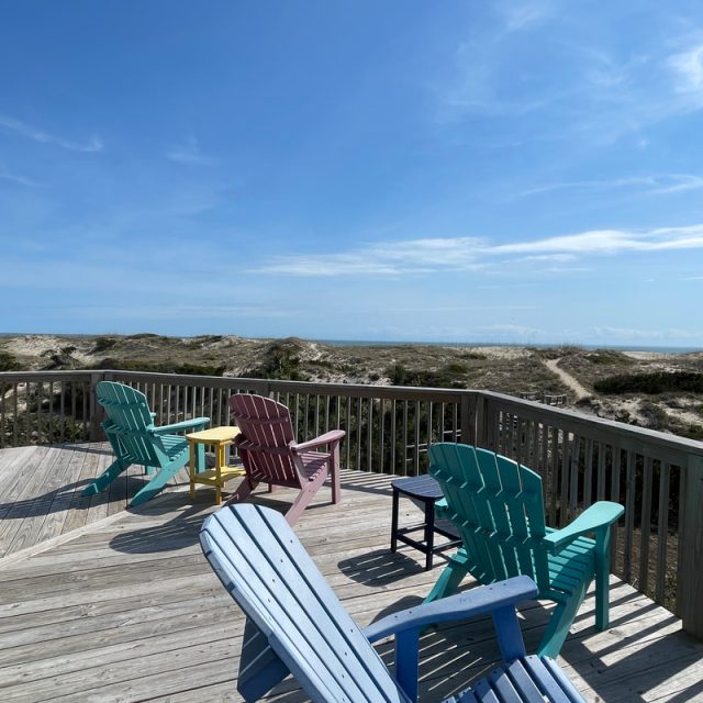 Five colorful Adirondack chairs on a wooden deck overlook sand dunes and blue sky at the beach.