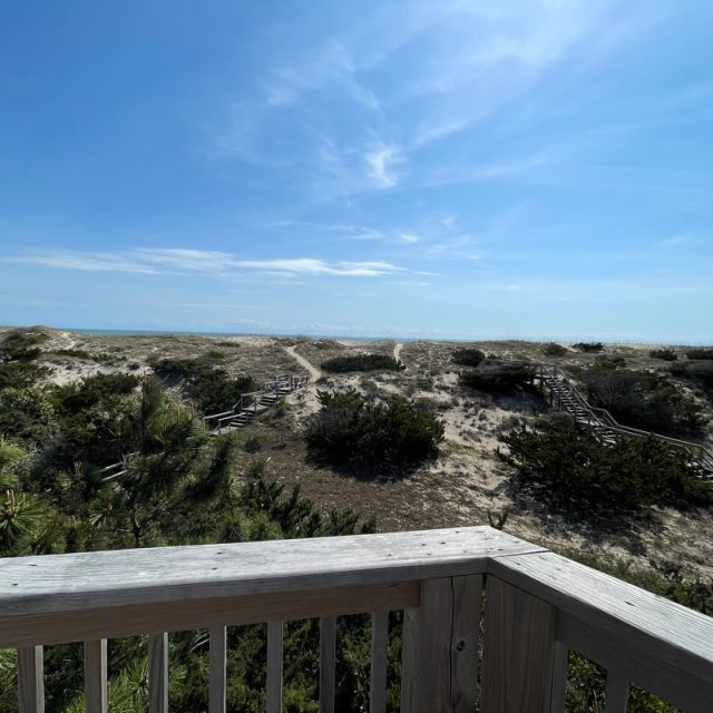 Wooden railing overlooks sandy dunes with scattered shrubs and a blue sky with wispy clouds in the background.