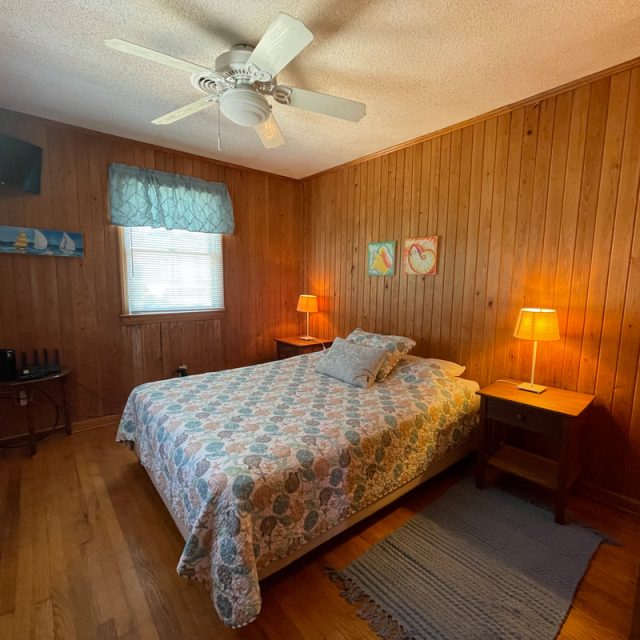 Wood-paneled bedroom with a double bed, two side tables with lamps, a ceiling fan, and wall art above the bed.