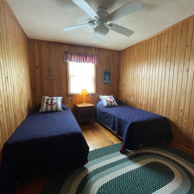 Wood-paneled bedroom with two twin beds, a window, a ceiling fan, and nautical-themed decor.