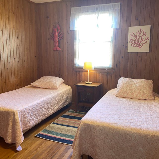Two twin beds with pink quilts in a wood-paneled room, a window with lamp, and sea-themed wall art.