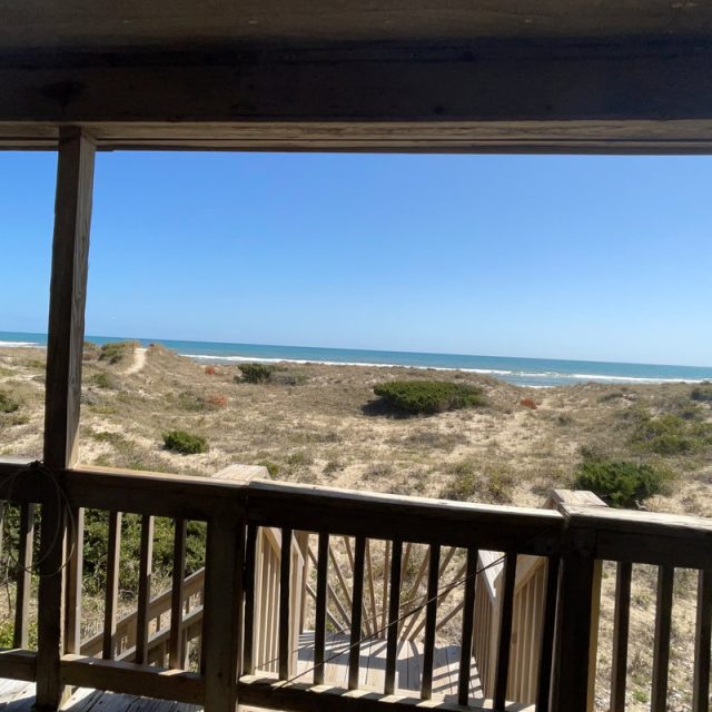 View of sandy dunes, shrubs, and blue ocean seen from a wooden balcony under a shaded roof on a clear day.