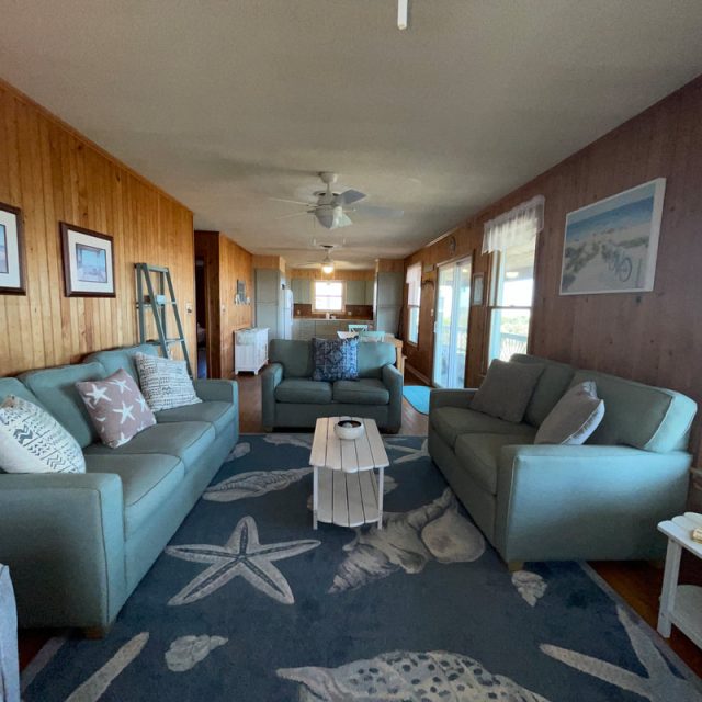 Living room with blue sofas, ocean-themed rug, wooden walls, framed art, and natural light from large windows.
