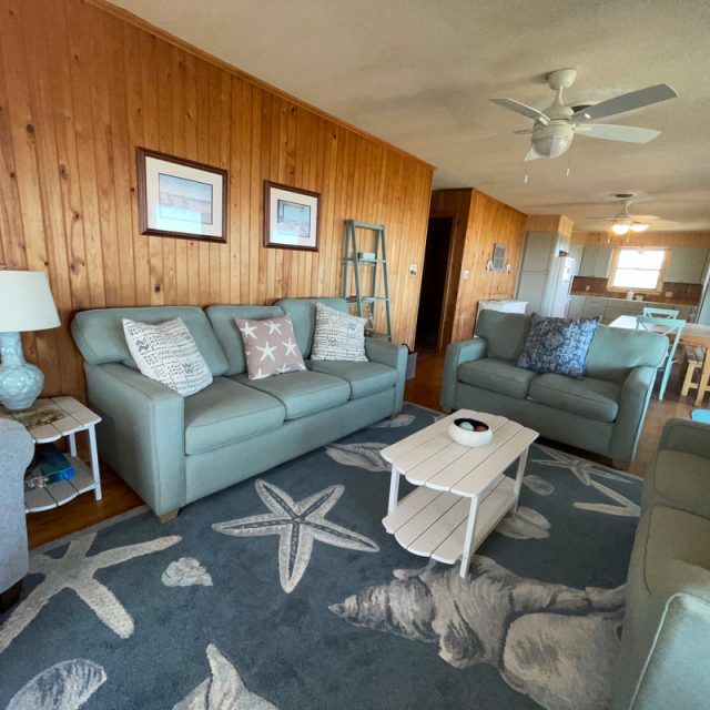 Cozy living room with light blue sofas, wood-paneled walls, and a starfish-themed rug, leading to dining area and kitchen.