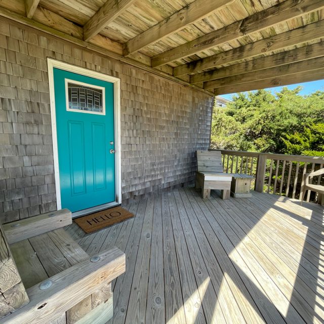 Wooden porch with a teal door, two bench seats, a small table, and a “Hello” doormat under partial sunlight.