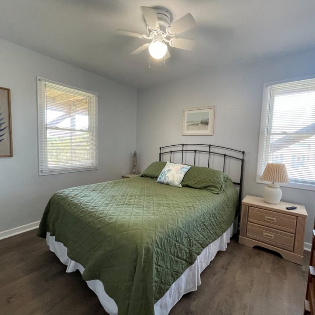 Bedroom with a green bedspread, two windows, dresser, nightstand with lamp, and simple wall art.