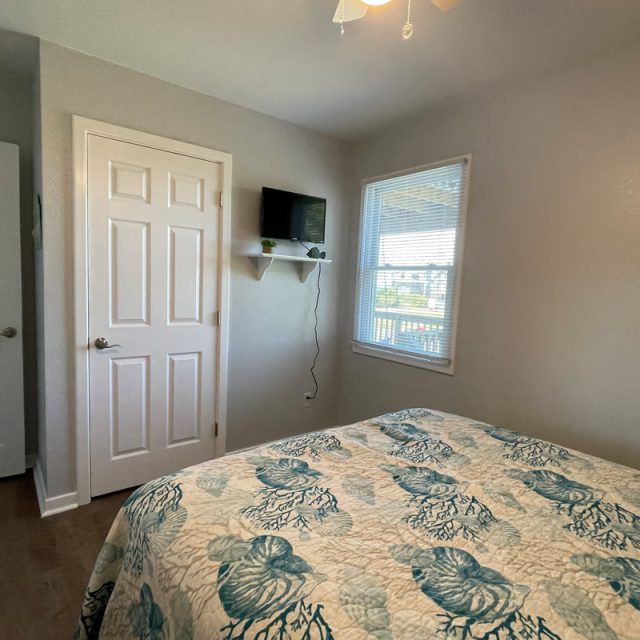 Small bedroom with a bed, patterned quilt, two closed doors, a wall-mounted TV, and a window with daylight.