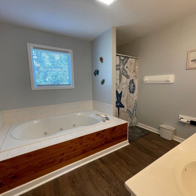 Bathroom with a large bathtub, double sinks, and a shower with a patterned curtain. Window and decor on the walls.