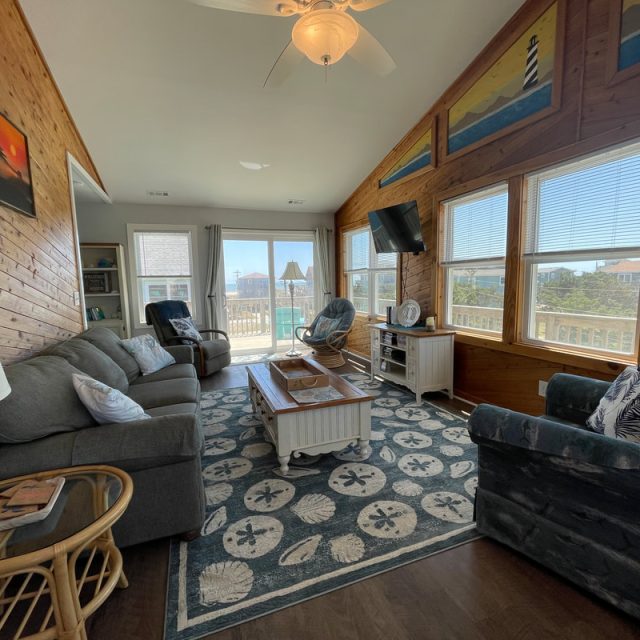 Living room with large windows, coastal decor, sofas, a coffee table, TV, and view of deck and houses outside.