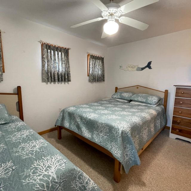 Bedroom with a double bed, a twin bed, wooden dresser, ceiling fan, and small windows with patterned curtains.
