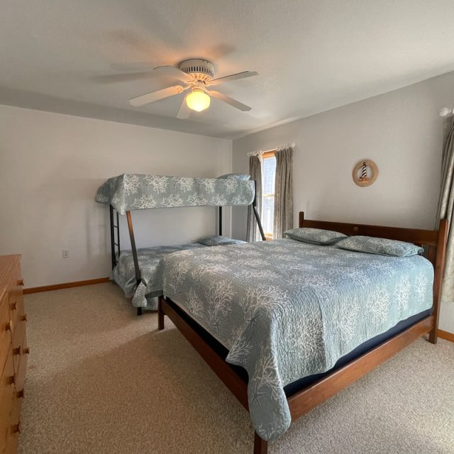 Bedroom with a double bed and bunk bed, light blue bedding, dresser, lamp, ceiling fan, and two windows with curtains.