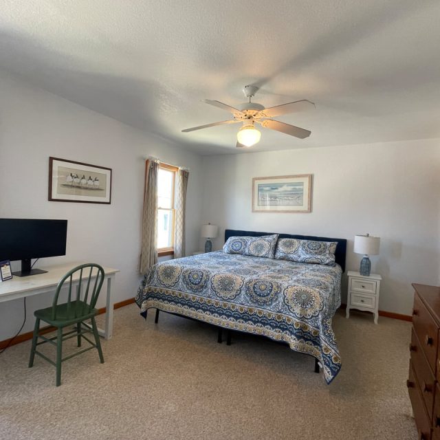 Bedroom with a bed, desk and chair, dresser, two windows with curtains, ceiling fan, and wall art.