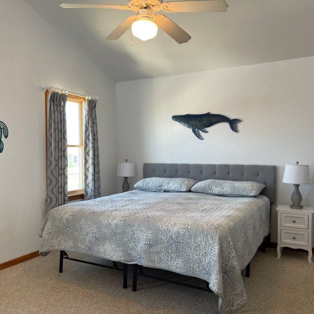 Bedroom with a king bed, two nightstands, sea-themed wall art, window with curtains, and a ceiling fan.