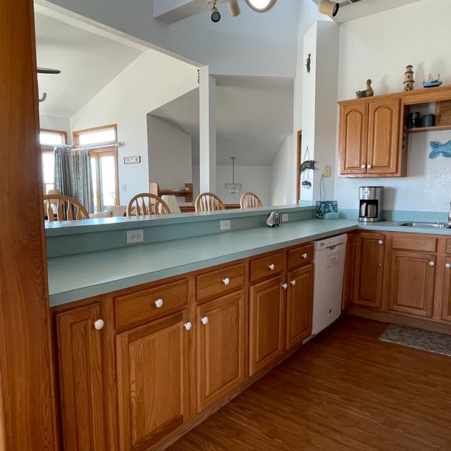 Kitchen with wooden cabinets, light blue countertops, a dishwasher, coffee maker, and dining area visible in the background.