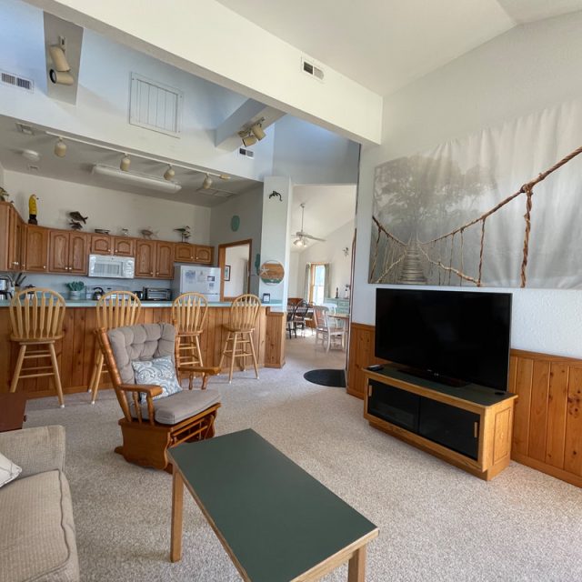 Living room with sofa, armchair, TV, and kitchen with wooden cabinets and barstools in an open floor plan.