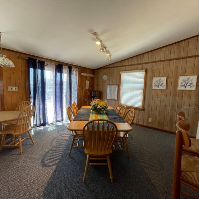 Dining room with wooden walls, two tables with chairs, large window with curtains, and a vase of flowers on the table.