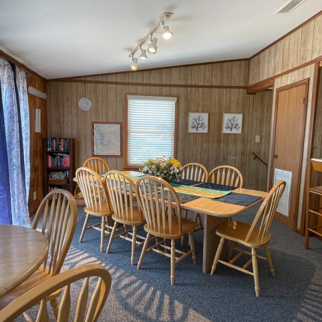 Dining area with a large wooden table, ten chairs, floral centerpiece, round table, and wood-paneled walls.