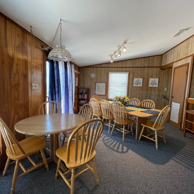 Dining room with wood-paneled walls, two tables, wooden chairs, large windows, and blue carpeted floor.