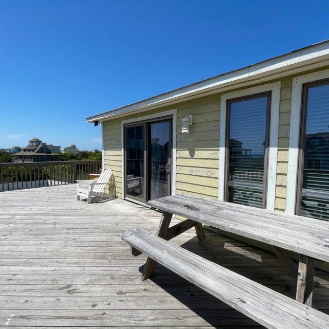 Wooden deck with picnic table and chairs outside a yellow house, overlooking nearby homes under a clear blue sky.