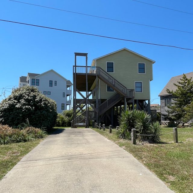 Light green beach house on stilts with exterior stairs, driveway, and grassy yard on a clear sunny day.