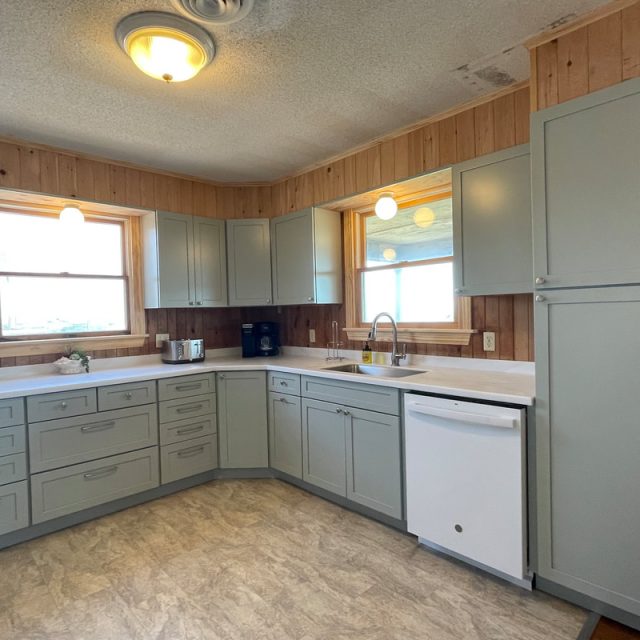 Kitchen with light green cabinets, wood panel walls, white countertops, and appliances under bright ceiling lights.