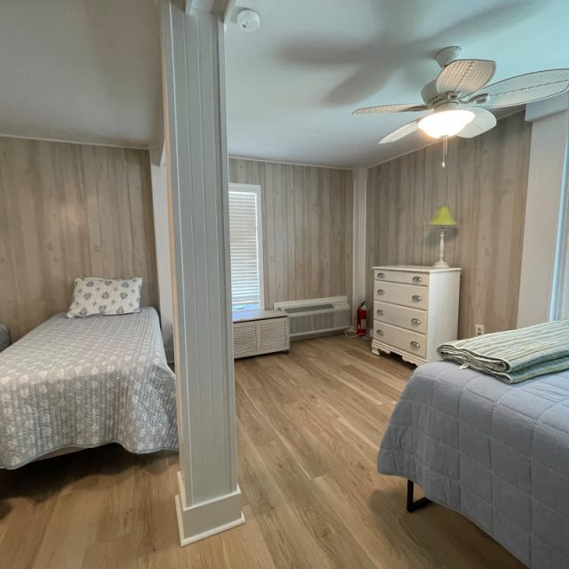 Bedroom with two twin beds, wood floors, a ceiling fan, dresser, and windows with blinds. Folded towels on one bed.