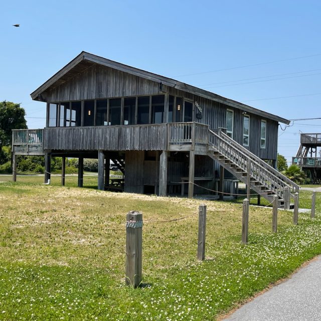 Wooden house on stilts with a front porch, surrounded by grass, under a clear blue sky.