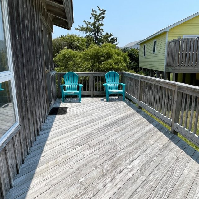 Two turquoise chairs sit on a wooden deck with railings, attached to a house, on a sunny day.