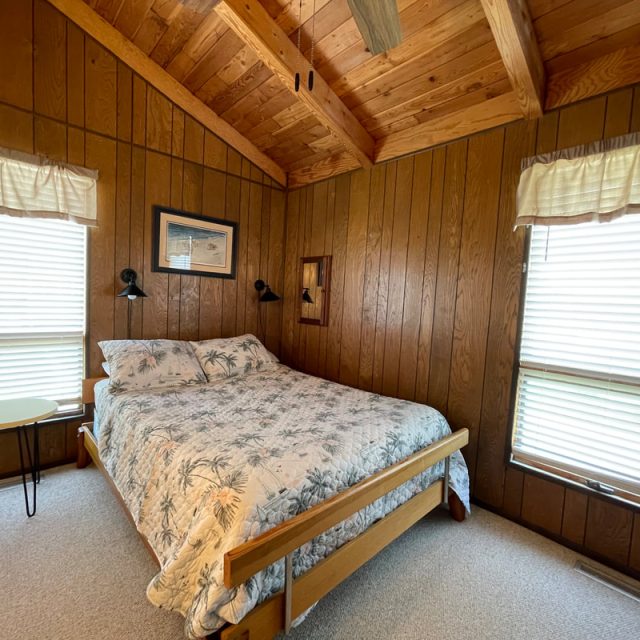 Wood-paneled bedroom with a double bed, two windows with blinds, and a small round table beside the bed.