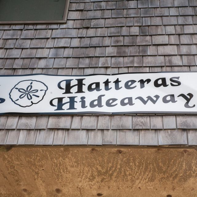 A wooden sign reads “Hatteras Hideaway” with a sand dollar illustration, mounted on a shingled wall.