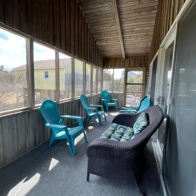 Screened porch with wicker loveseat, blue plastic chairs, and view of a yellow house outside.