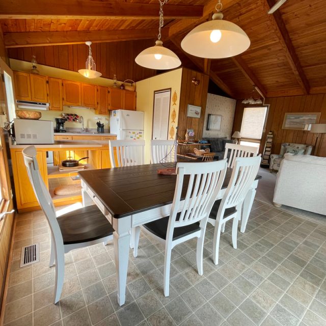 A wooden dining table with six white chairs sits in an open kitchen and living room with wood-paneled walls and ceiling.