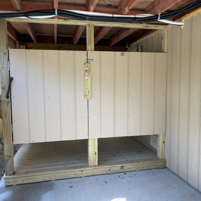 Outdoor wooden enclosure with a lock under a raised roof, adjacent to a fence and siding, on a concrete pad.