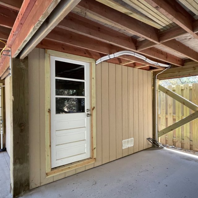 A wooden door with three glass panes set in a tan wall under a covered patio next to a wooden fence.