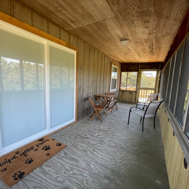 Screened-in porch with sliding glass door, small table and chairs, metal chair, and a welcome mat on concrete floor.