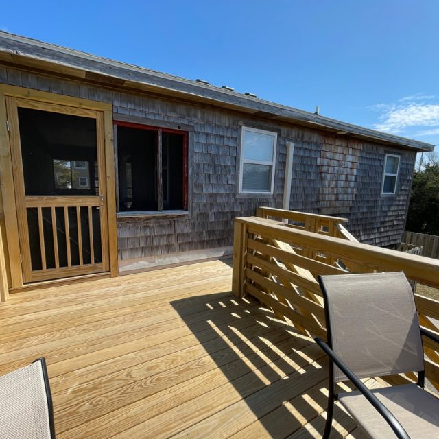 Wooden deck with chairs facing a shingled house, under a clear blue sky.