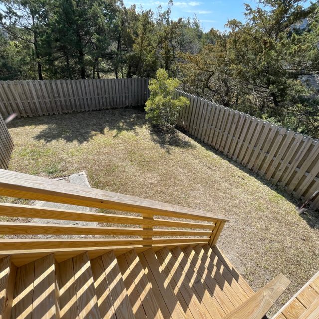 View from wooden stairs overlooking a fenced backyard with grass, trees, and sunlight casting shadows.