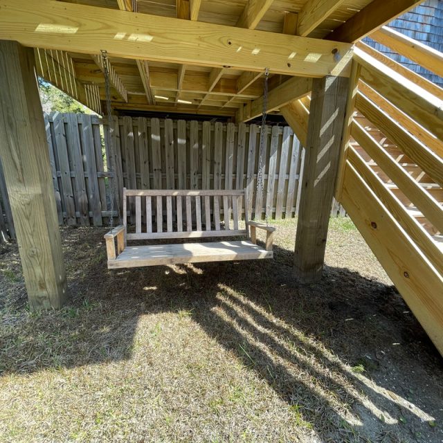 Wooden porch swing hanging under a deck next to a wooden staircase and a fenced backyard area on a sunny day.