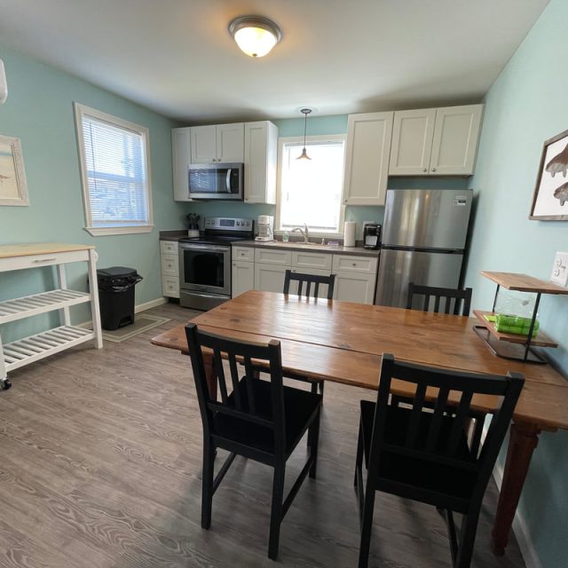 Small kitchen with white cabinets, stainless appliances, wooden table with four black chairs, and light blue walls.