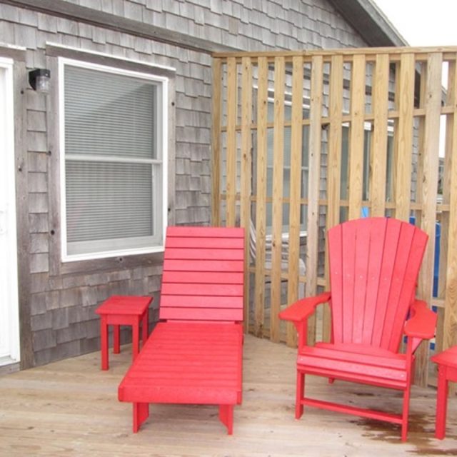 Two red wooden chairs and two small red tables on a wooden deck next to a house with gray siding and window blinds.
