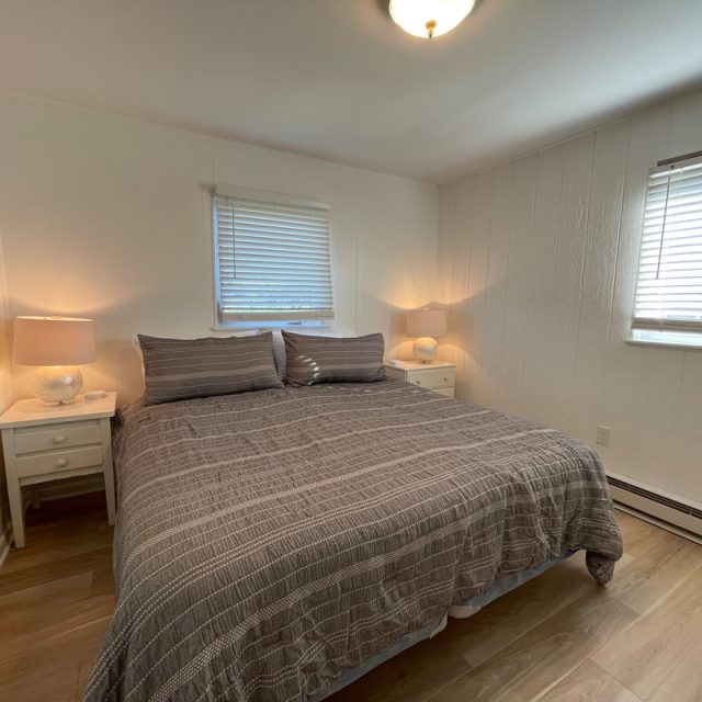 Bedroom with a king-size bed, two nightstands with lamps, wood floor, and two windows with blinds.