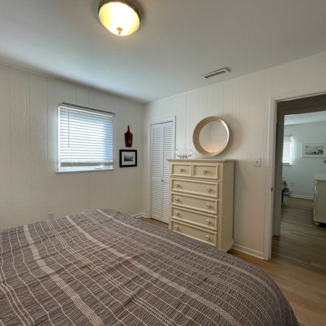 Bedroom with a striped bedspread, dresser with a round mirror, and a window with blinds; door open to another room.