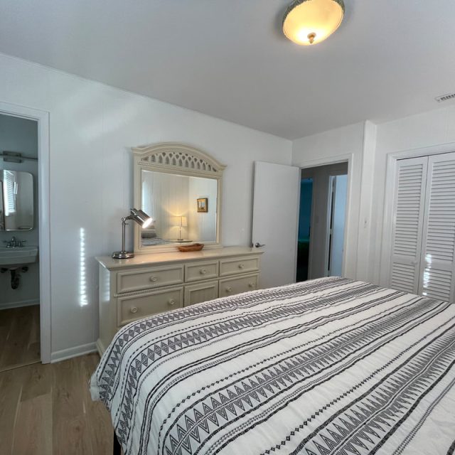 Bedroom with striped bedspread, dresser with mirror, lamp, and open door leading to a small bathroom.
